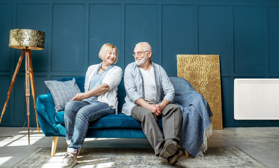 Couple senior souriant assis sur un canapé bleu dans un salon moderne, avec un radiateur électrique mural en arrière-plan.